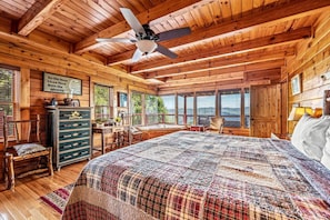 Een knus slaapkamer in chaletstijl met een groot bed, houten wanden, een plafondventilator en uitzicht op de natuur door grote ramen.