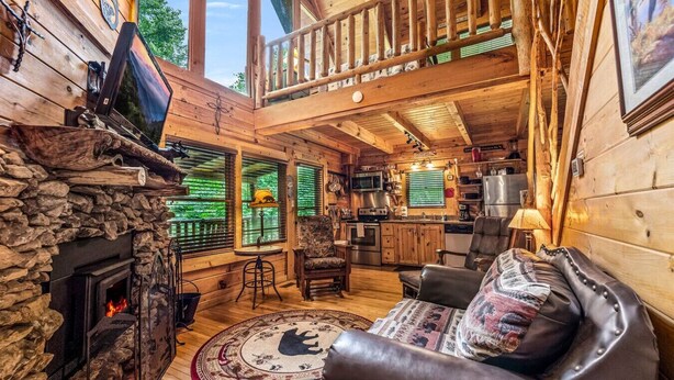 A cozy cabin interior with a stone fireplace, wooden walls, a balcony, and a kitchen area. - 2BR Resort Cabin w/Hot Tub + Fireplace + Privacy! (Sevierville)