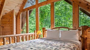 A wooden cabin bedroom with a large bed, a wooden headboard, and a view of lush greenery through large windows. - 2BR Resort Cabin w/Hot Tub + Fireplace + Privacy! (Sevierville)