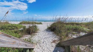 Un sentier de sable qui longe la plage et mène à l’océan, bordé de végétation.