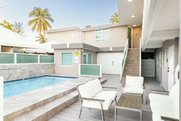 A swimming pool with steps leading to a covered area, white lounge chairs, and a staircase leading to an upper level.