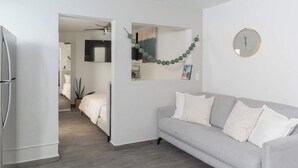 En moderne stue med en grå sofa, et rundt vægur og en gang der fører til et soveværelse.
