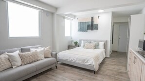 Une chambre moderne avec un canapé gris, un lit avec une literie blanche, une table de chevet et une petite table avec une lampe.
