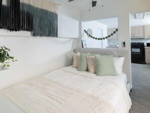 A bedroom with a bed, pillows, a bedside plant, and a view into the kitchen. - AQUA Terrazas 3 · Seaside Fun & Dining with Pool (Dorado)