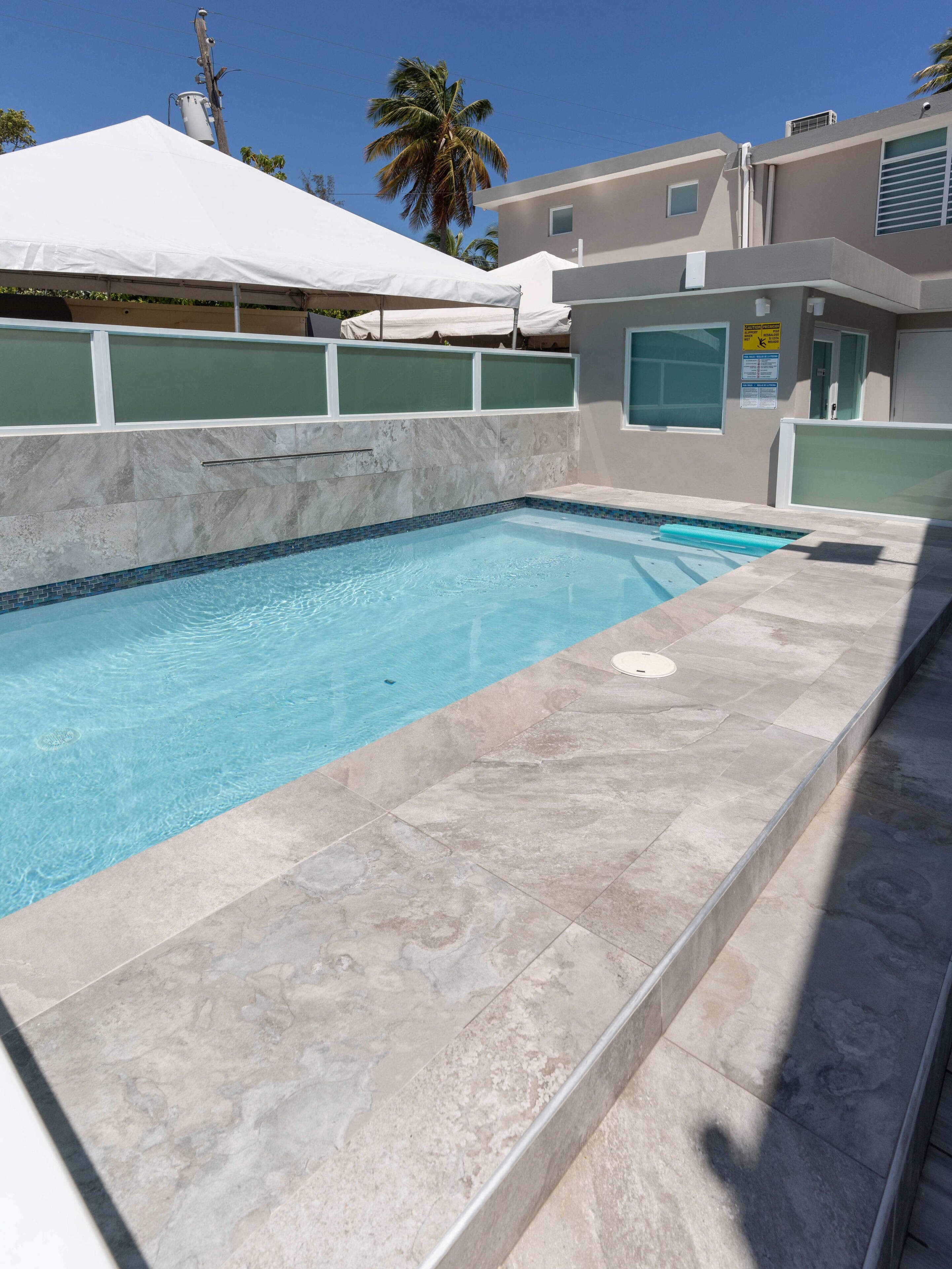 Piscina com deck ladrilhado, toldo branco e palmeira ao fundo.
