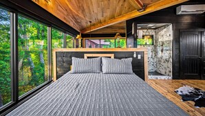 Un dormitorio con un ventanal grande, techo de madera y una cama con ropa de cama gris.