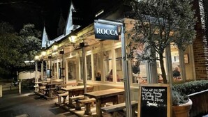 Un restaurante llamado Roccaa con terraza y un cartel que indica el horario de apertura.