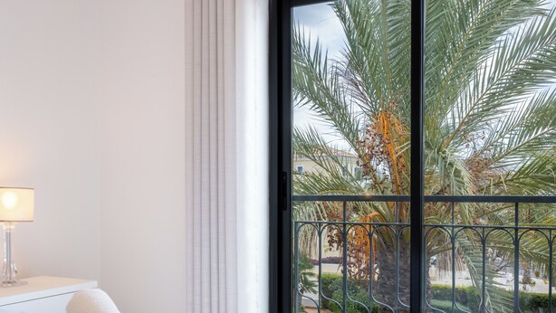 A hotel room with a bed, a nightstand with a lamp, and a view of a swimming pool and palm trees through a window with a metal railing.
