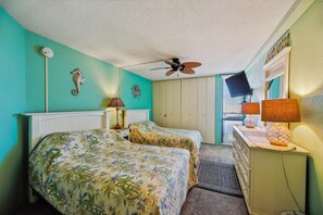A room with two beds, a ceiling fan, a desk with a lamp, and a window with blinds.