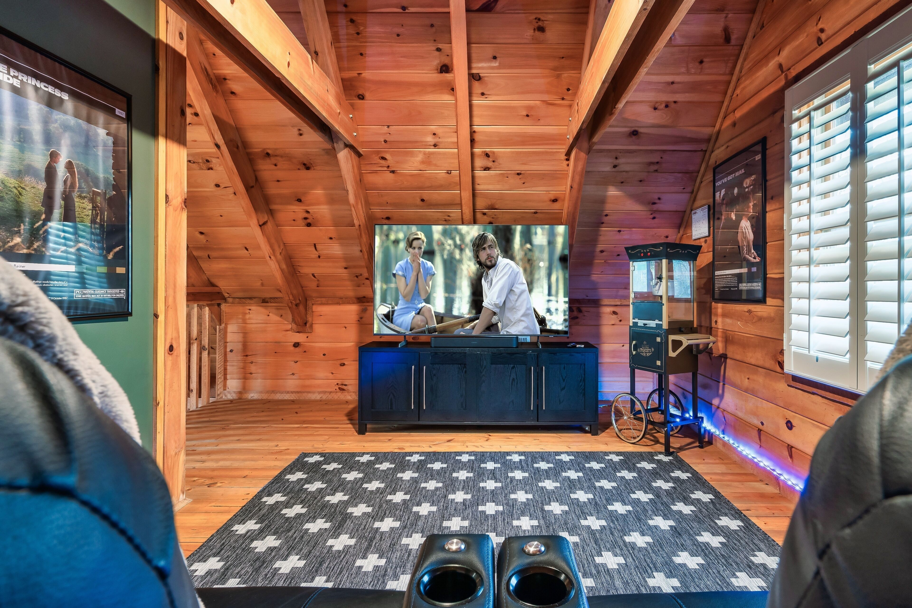 Ein gemütliches Blockhaus-Interieur mit Holzbalkendecke, einem an der Wand befestigten Fernseher, einem gemusterten Teppich und zwei Lautsprechern.