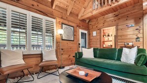 Un interior de cabaña acogedor con un sofá verde, dos sillones de cuero, una mesa redonda de centro y un estante de madera con varios objetos.