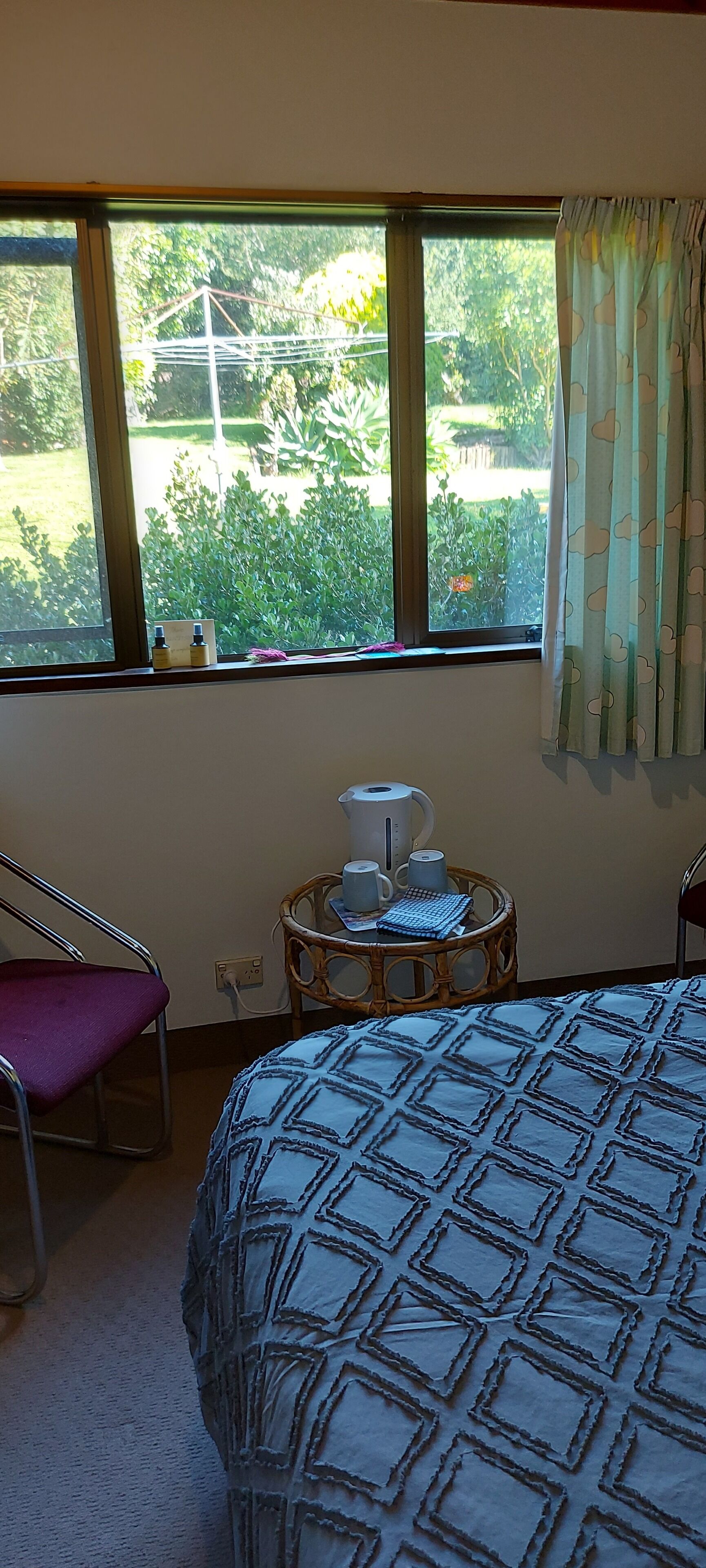 A bedroom with a bed, a chair, a small table with a kettle and cups, and a window with a view of greenery.