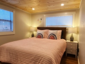 Un dormitorio con techo de madera, dos lámparas de noche, una cama con colcha rosa y dos ventanas con persianas.
