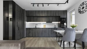 Eine moderne Küche mit dunklen Schränken, einer zentralen Kochinsel und einem Essbereich mit weißem Tisch und grauen Stühlen.