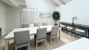 A modern dining room with a high ceiling, a skylight, a dining table with six chairs, a potted plant, and a painting on the wall.