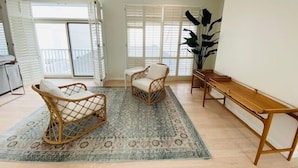 A spacious living area with a large window, a wooden bench, two wicker chairs, and a patterned rug.