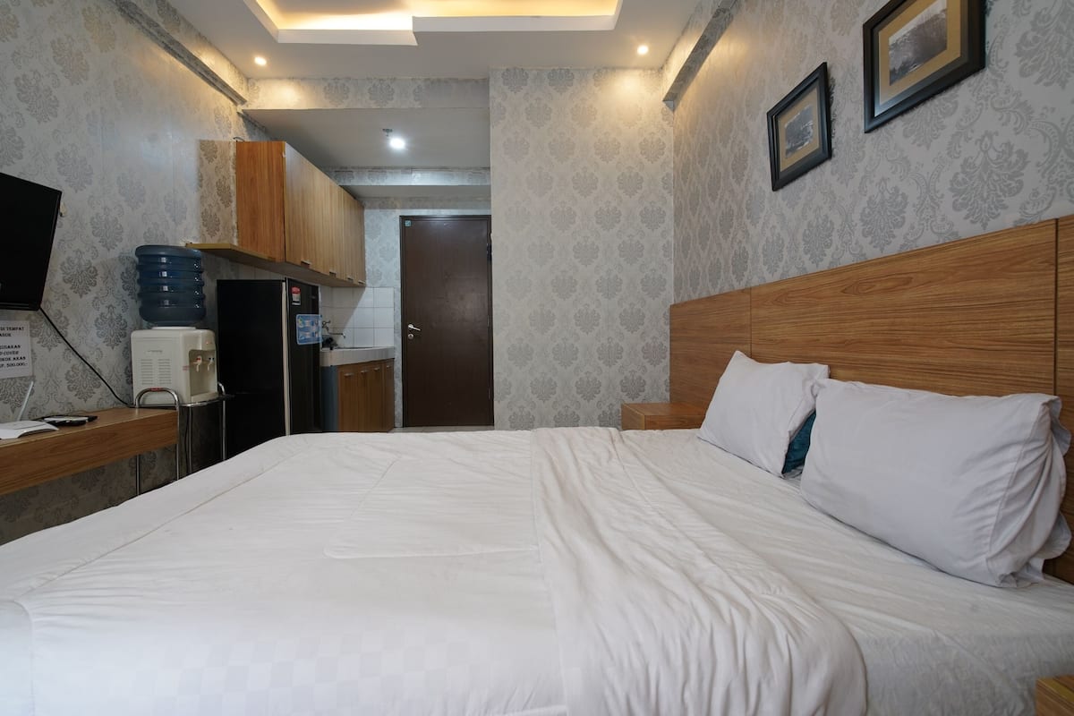 Une chambre d’hôtel avec un grand lit, une tête de lit en bois et une petite table de chevet. Elle comprend une kitchenette équipée d’un réfrigérateur et d’un distributeur d’eau. Les murs sont recouverts d’un papier peint à motifs et une porte donne accès à un autre espace.