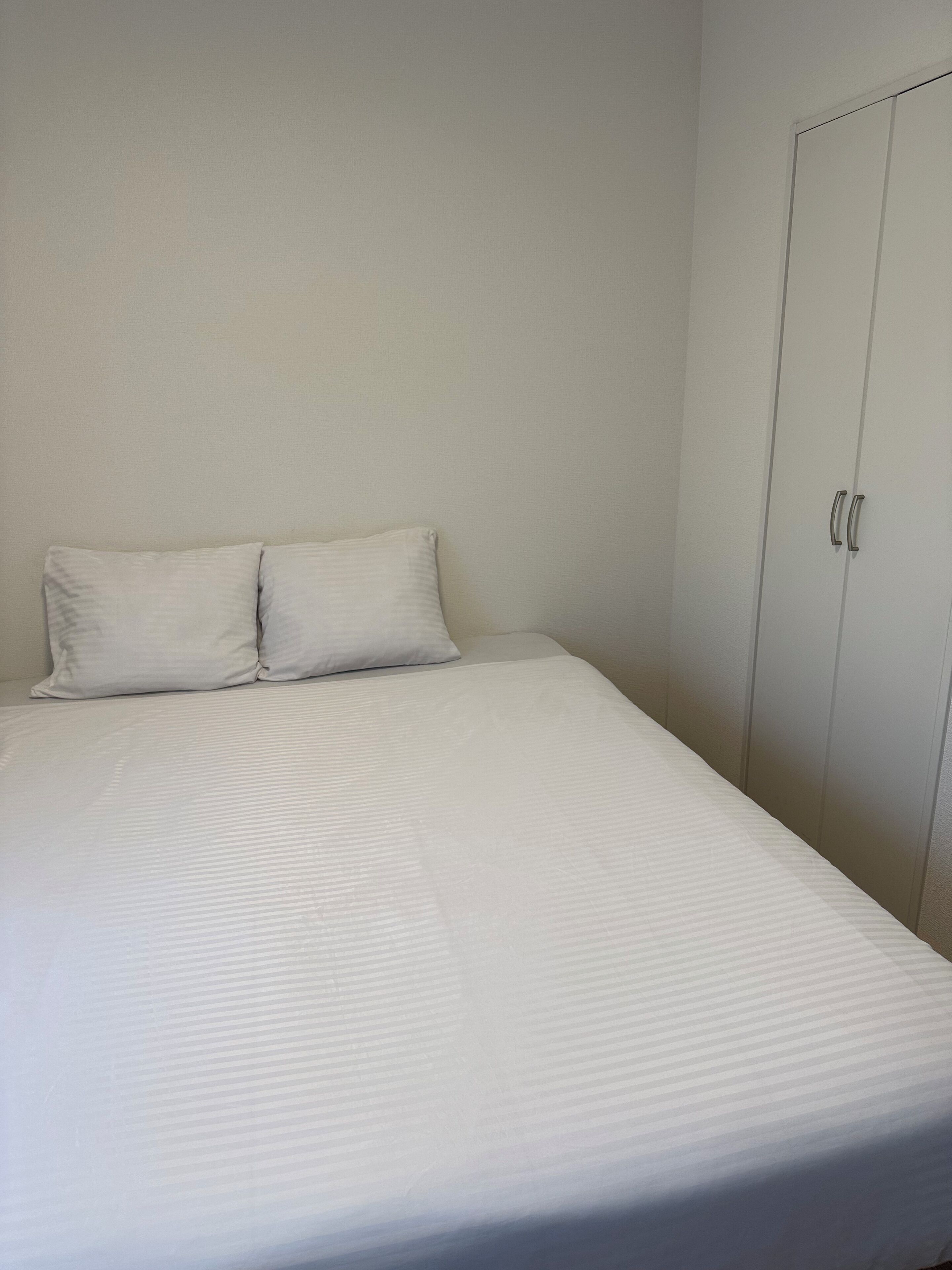 A single bed with a white mattress and two pillows, a white wardrobe with two doors, and a plain wall.
