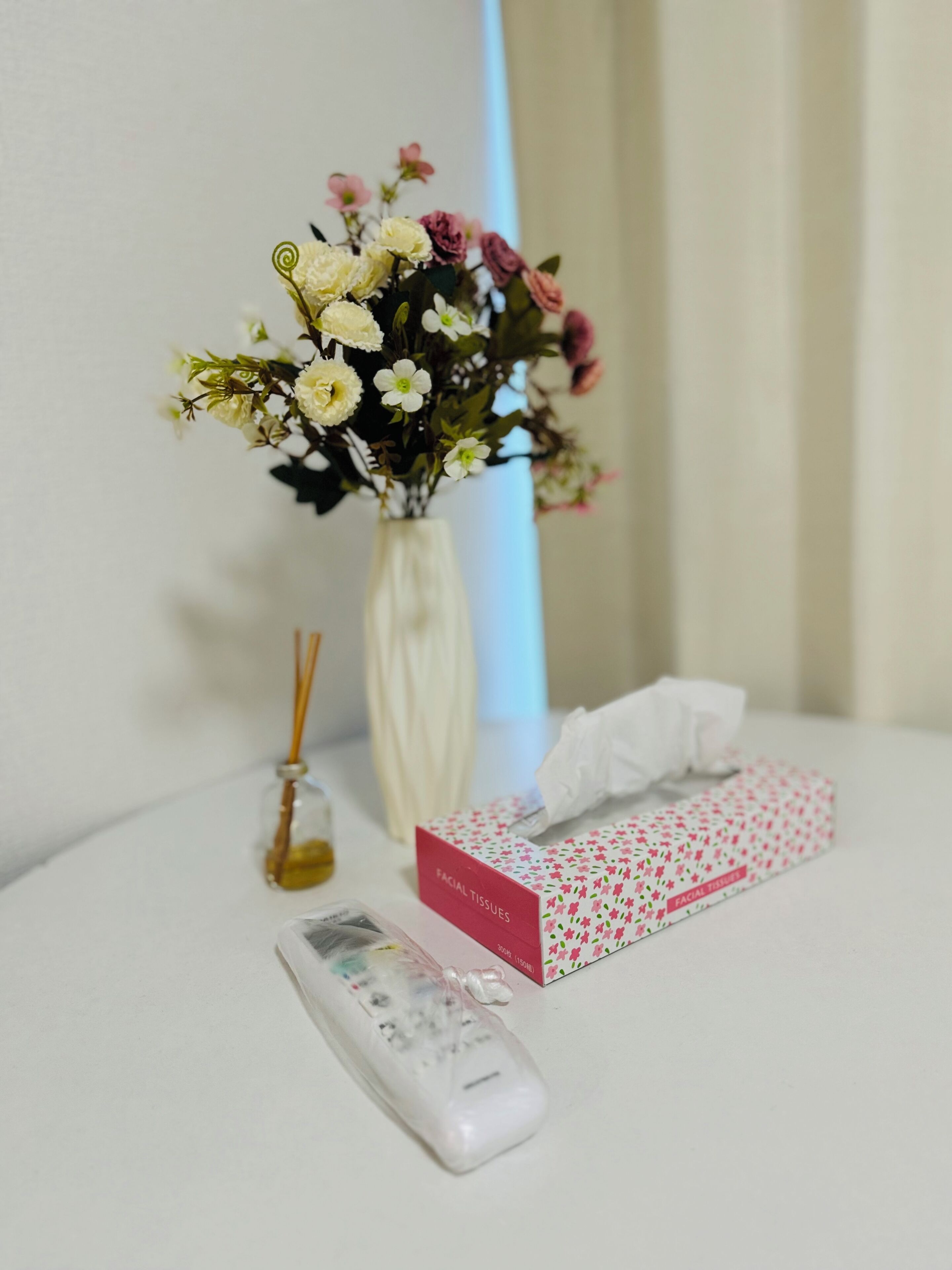 A white vase with flowers, a box of facial tissues, and a remote control on a white table.