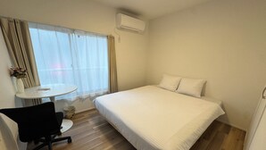 Une petite chambre d’hôtel, bien rangée, avec un lit simple, une petite table ronde, une chaise et une fenêtre avec des rideaux.