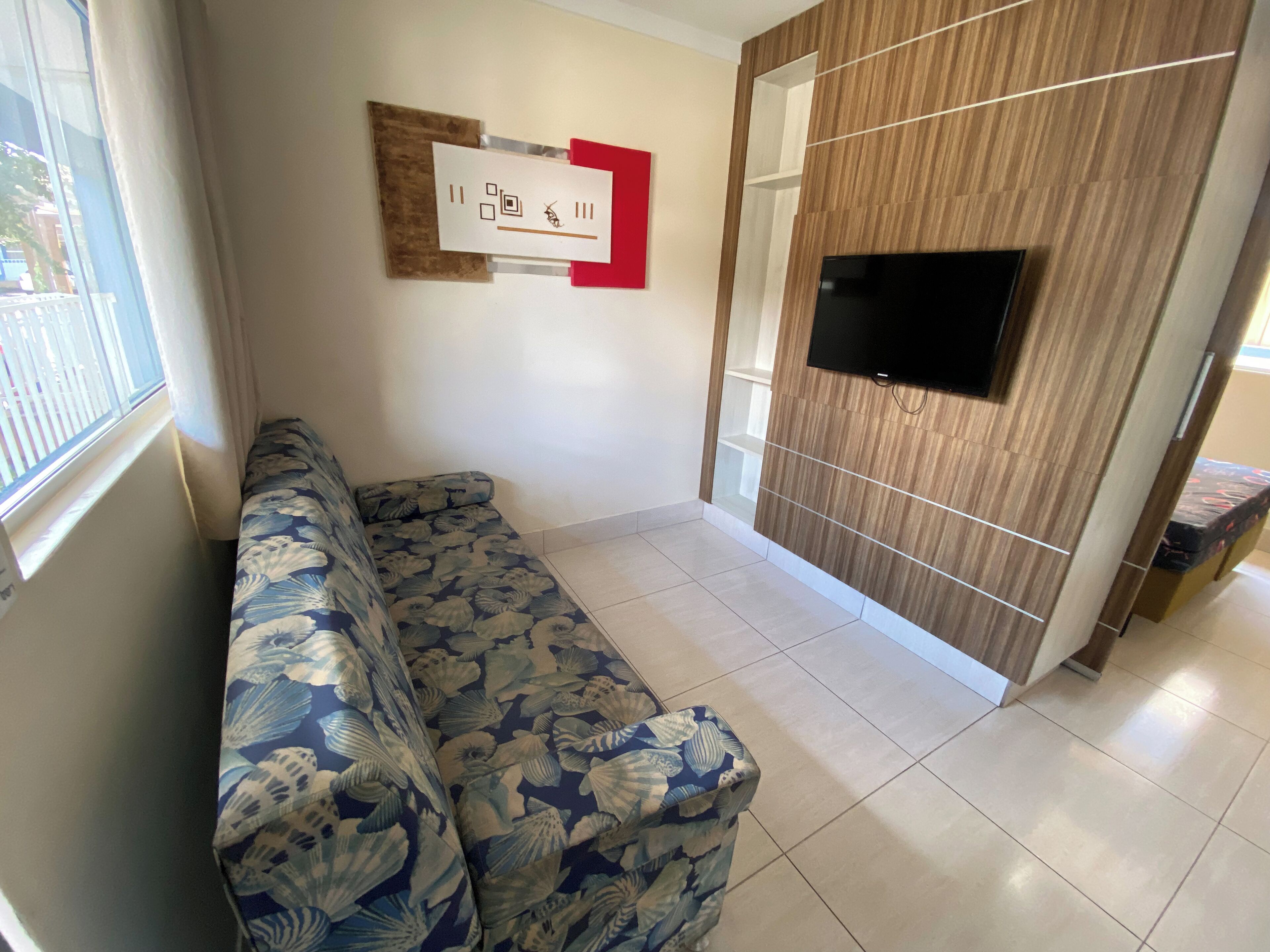 A living room with a sofa, a TV mounted on a wooden panel wall, and a window with curtains.