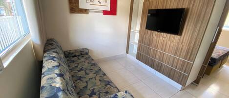 A living room with a sofa, a TV mounted on a wooden panel wall, and a window with curtains.
