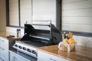 A kitchen with a built-in grill, a countertop with a basket of bread, and a window with blinds. - Airlie Nights (Airlie Beach)
