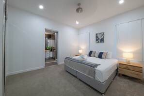 A modern bedroom with a large bed, bedside lamps, a small bedside table, and a door leading to a bathroom. - Airlie Nights (Airlie Beach)