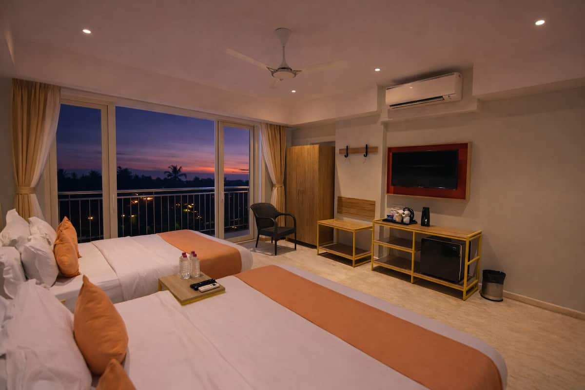 A hotel room with two beds, a TV, a desk, and a balcony with a view of the sunset.