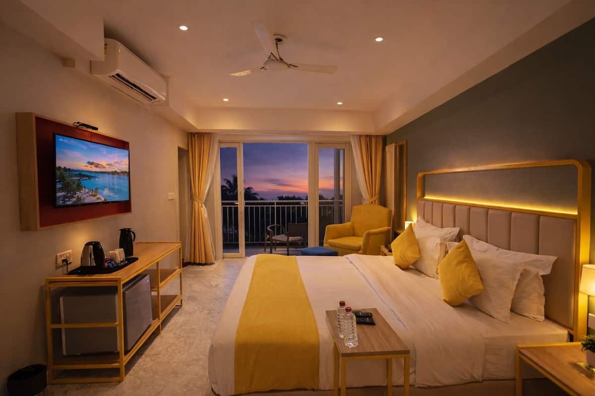 A hotel room with a bed, a desk, a chair, a TV, and a balcony with a view of the sunset.