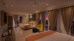 Une chambre d’hôtel avec un grand lit, un plus petit lit, une chaise, un bureau et une vue sur la ville au crépuscule.
