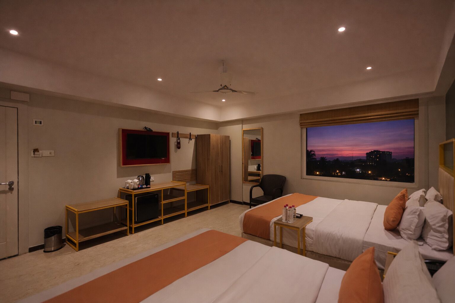 A hotel room with two beds, a desk, a TV, a wardrobe, and a window with a city view.