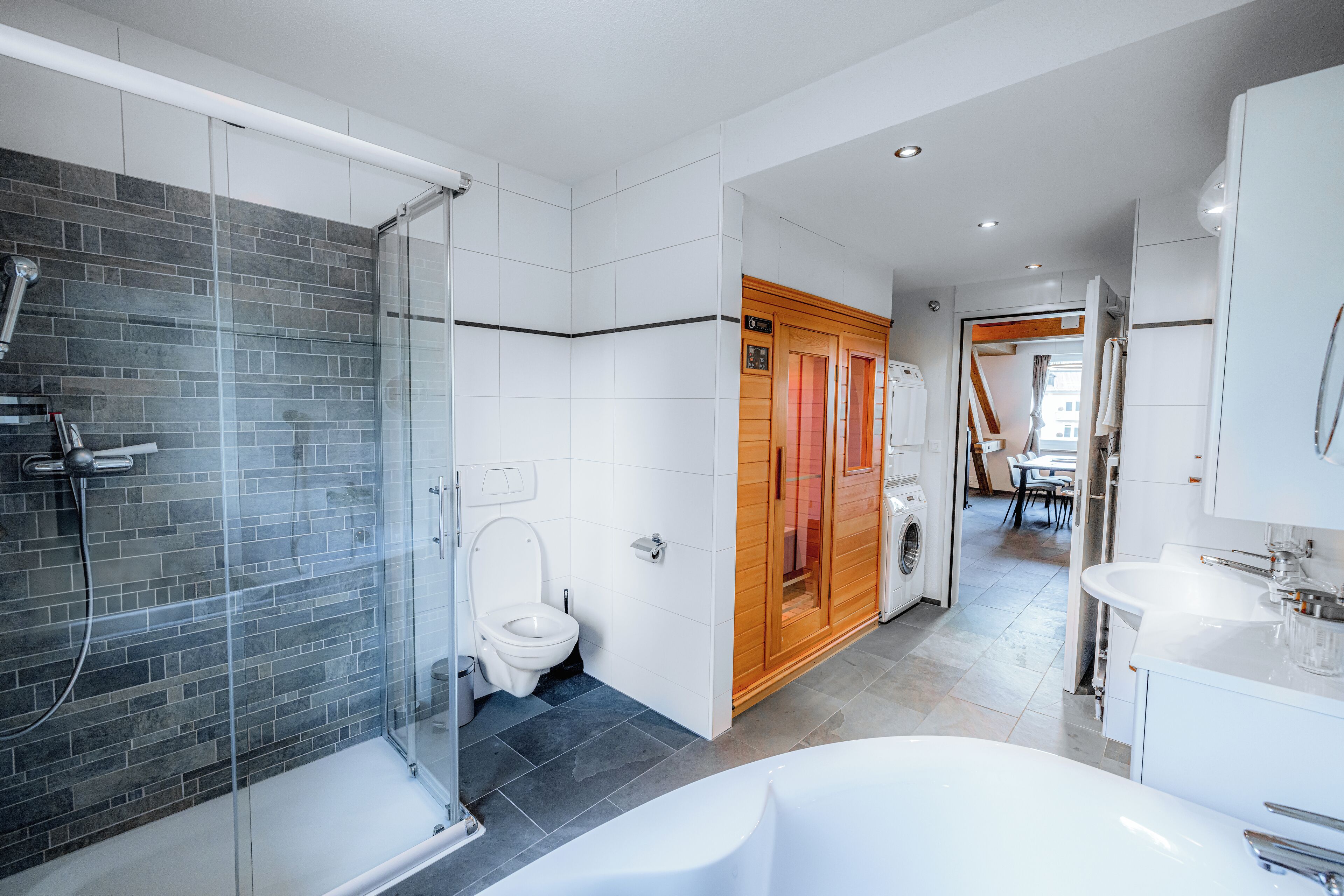 A modern bathroom with a glass-enclosed shower, a white bathtub, a wooden vanity, and a visible laundry area.