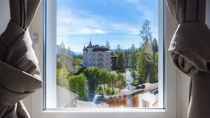 A view through a window with curtains, featuring a building with a tower, surrounded by trees and mountains.
