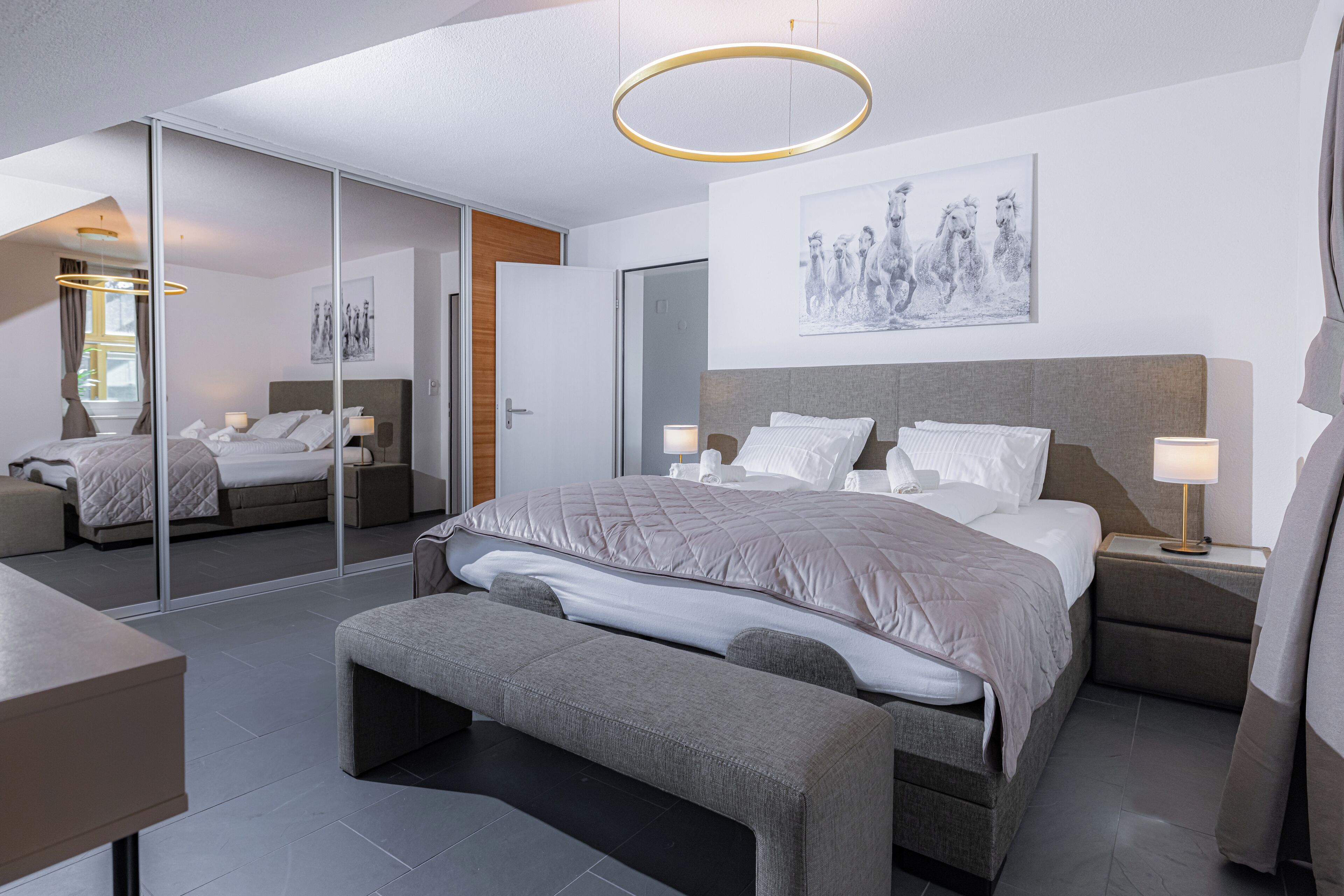 Una habitación de hotel moderna con una cama grande, un banco, una mesita de noche con lámpara, un espejo y una obra de arte colgada en la pared.