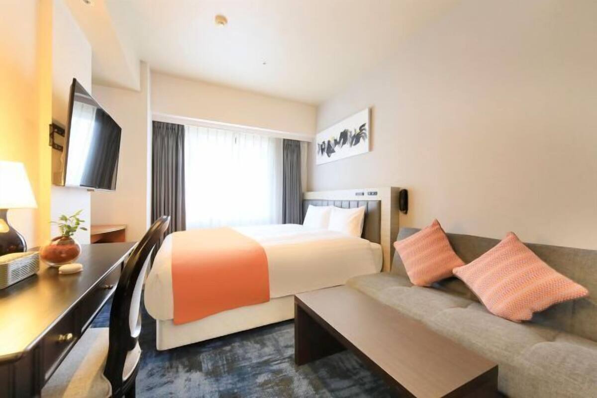 Habitación de hotel con cama, sofá, escritorio, televisor y ventana con cortinas.