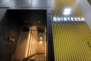L’entrée d’un hôtel, avec une enseigne portant le nom « QUINTESSA » et un design moderne caractérisé par des lignes verticales.