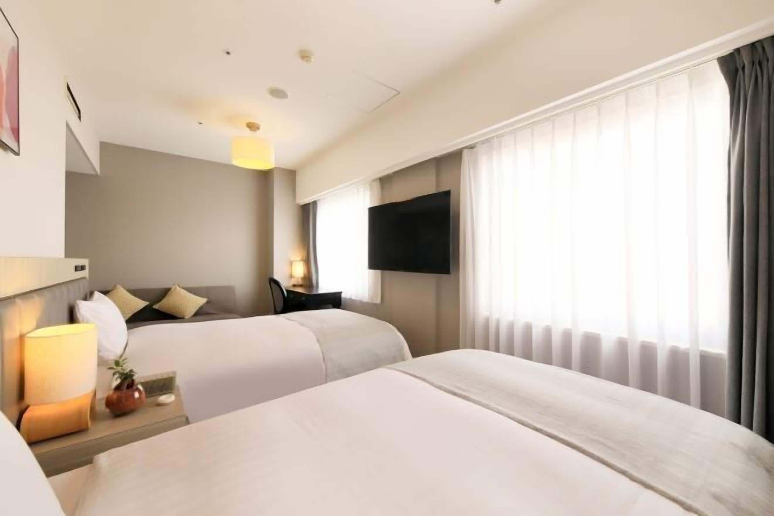 A hotel room with two beds, a flat-screen TV mounted on the wall, a bedside table with a lamp, and a window with curtains.