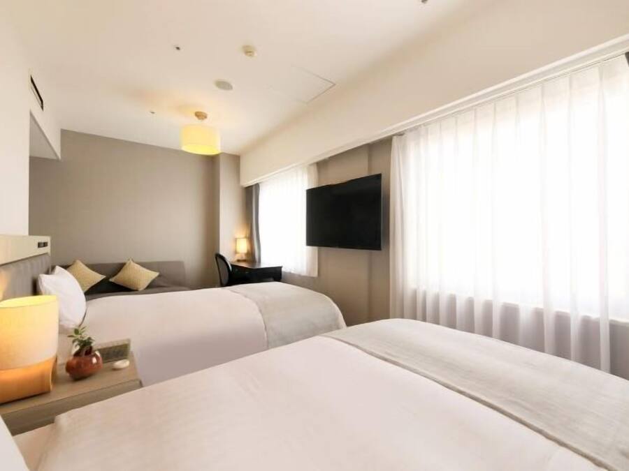 Ein Hotelzimmer mit zwei Betten, einem wandmontierten Flachbildfernseher, einem Nachttisch mit Lampe und einem Fenster mit Vorhängen.