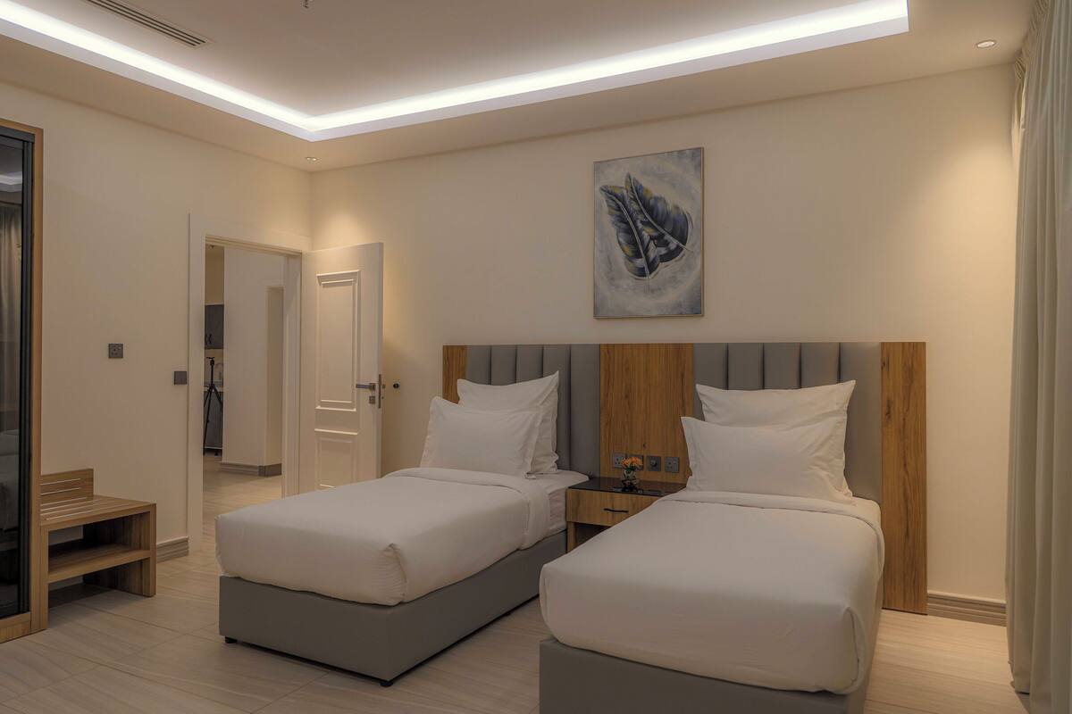 A hotel room with two beds, a wooden headboard, a nightstand, a bench, and a painting on the wall.