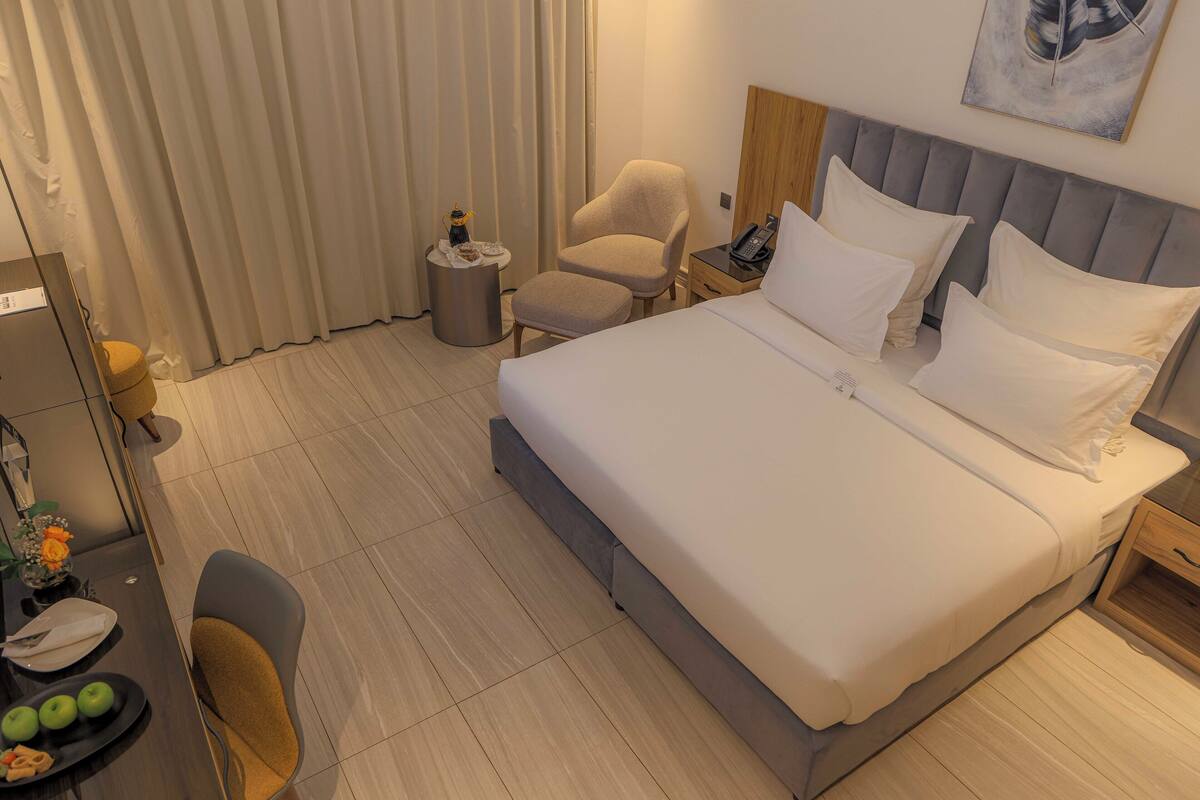 A hotel room with a large bed, a chair, a small table with a lamp, and a desk with a computer and a plate of fruit.
