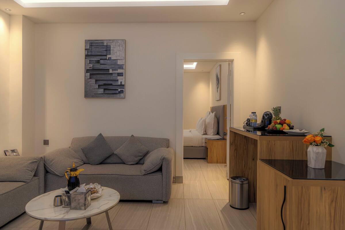 A modern living room with a grey sofa, a coffee table, and a kitchen area with a wooden counter and a fruit bowl.