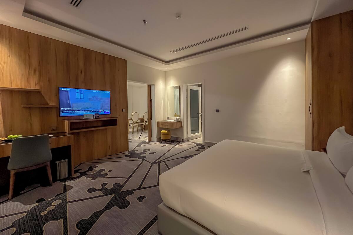 A hotel room with a bed, a desk with a computer, a television, and a bathroom visible through an open door.