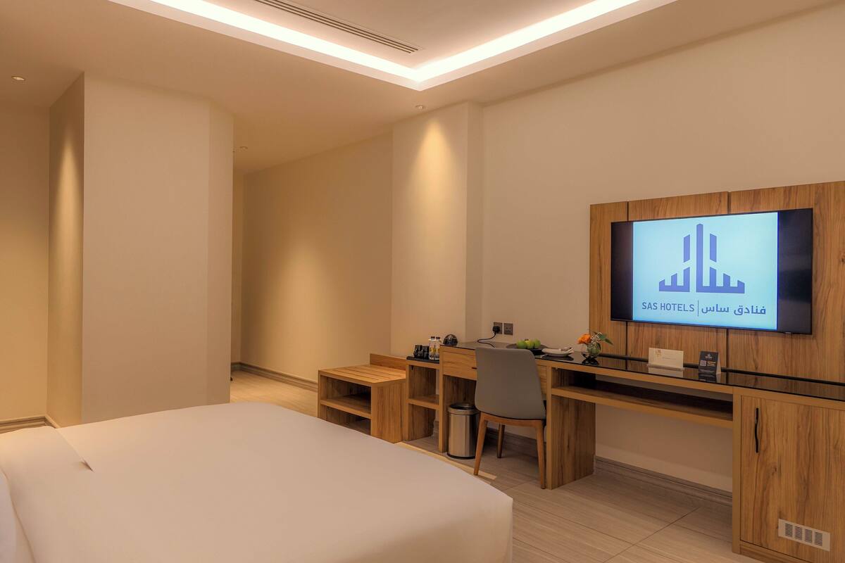A hotel room with a bed, a desk, a chair, a TV mounted on the wall, and a trash bin.