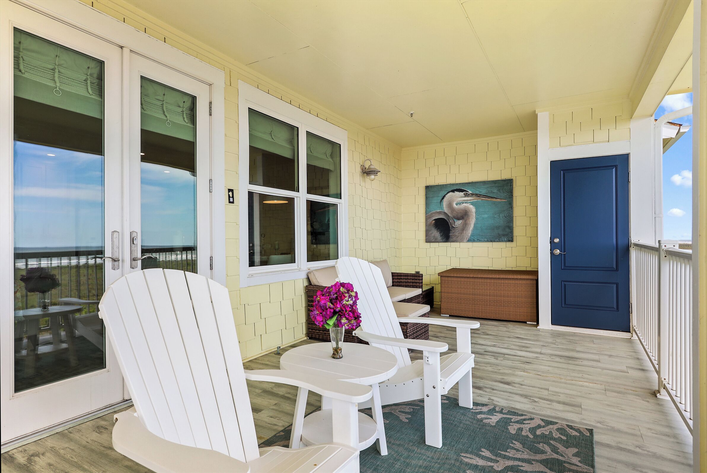 En overdækket veranda med hvide Adirondack-stole, et bord med en vase blomster, et maleri af en svane og en blå dør.