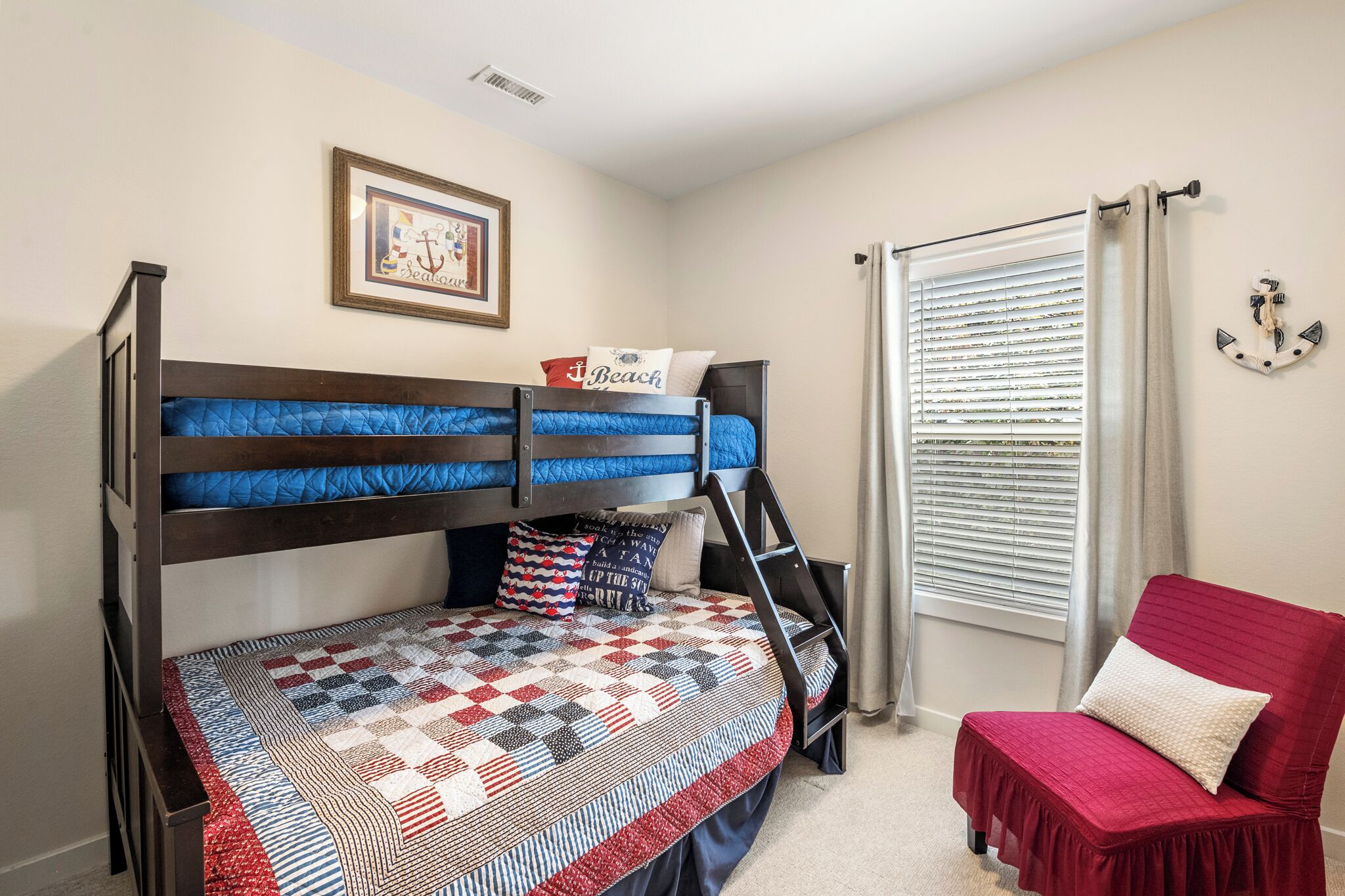 A bunk bed with a quilted headboard, a red chair, a window with blinds, and a framed picture on the wall.