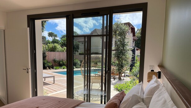 Une chambre dotée d’une grande porte-fenêtre coulissante donnant sur un jardin avec une piscine et du mobilier d’extérieur.