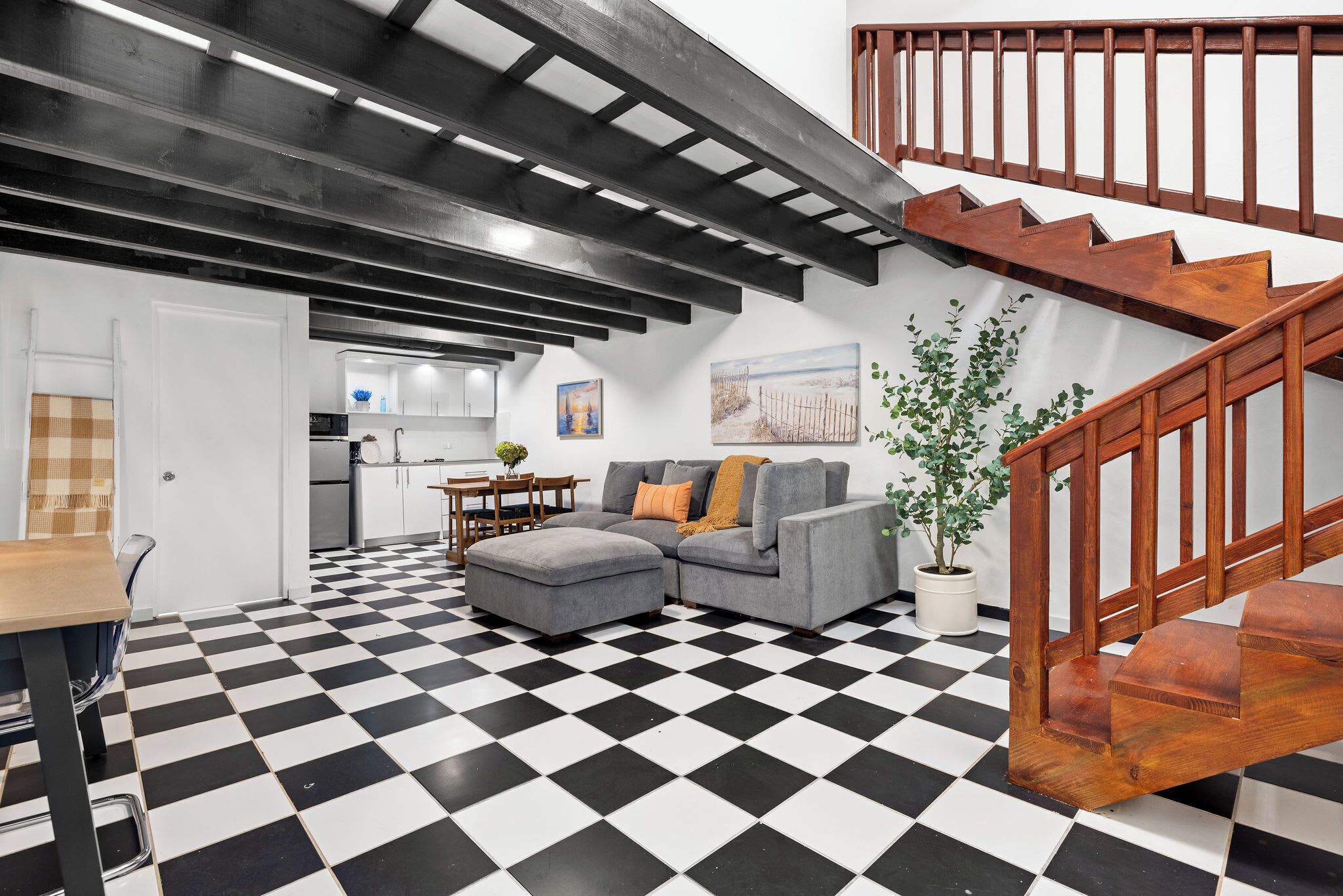 A living room with a checkered floor, a gray sofa, a wooden staircase, and a kitchen in the background.