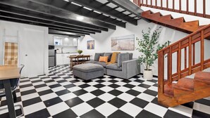 Ein Wohnzimmer mit Schachbrettmuster-Boden, einem grauen Sofa, einer Holztreppe und einer Küche im Hintergrund.