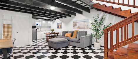A living room with a checkered floor, a gray sofa, a wooden staircase, and a kitchen in the background.